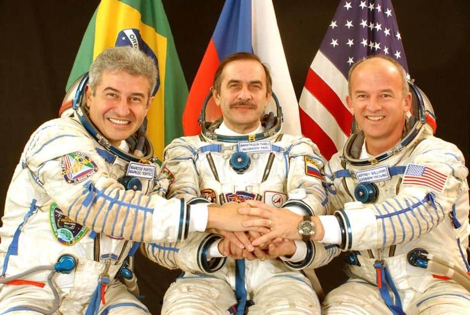 Da esquerda para a direita, o astronauta brasileiro Marcos Pontes, o cosmonauta russo Pavel Vinogradov e o astronauta norte-americano Jeffrey Williams, todos usando trajes espaciais. Atrás deles, as bandeiras do Brasil, Rússia e Estados Unidos. Eles estão dando as mãos, simbolizando união. Foto da NASA.