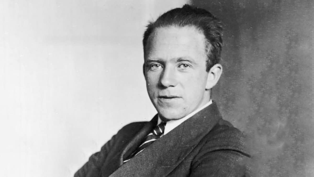 Werner Heisenberg, usando um terno preto e uma gravata listrada preta e branca. Ele tem um olhar sério e focado, com olhos intensos que refletem a profundidade de suas descobertas científicas. Sua expressão transmite a confiança de um dos principais pioneiros da mecânica quântica. O fundo é simples e neutro, destacando sua postura refinada e seu estilo clássico. Heisenberg, conhecido por sua formulação do Princípio da Incerteza, é retratado com um ar de autoridade intelectual.