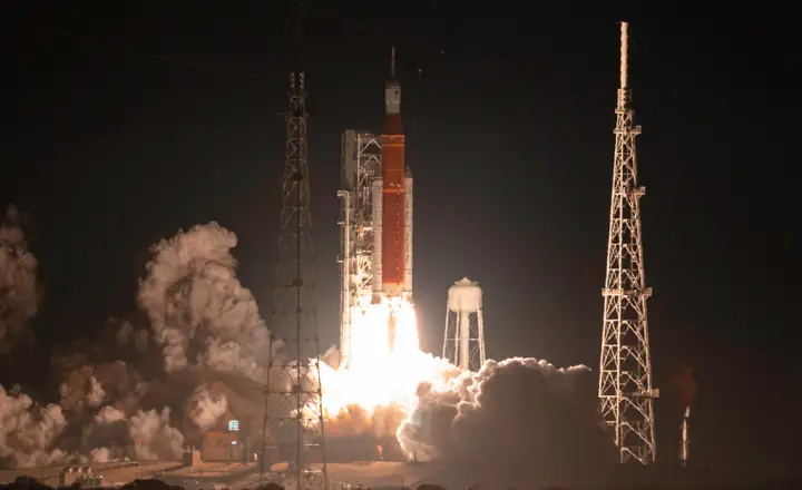 Lançamento do foguete SLS durante a Etapa 1 do Programa Artemis da NASA, mostrando o foguete laranja com boosters auxiliares brancos, a torre de lançamento ao fundo, e um clarão intenso com fumaça densa marcando a decolagem.