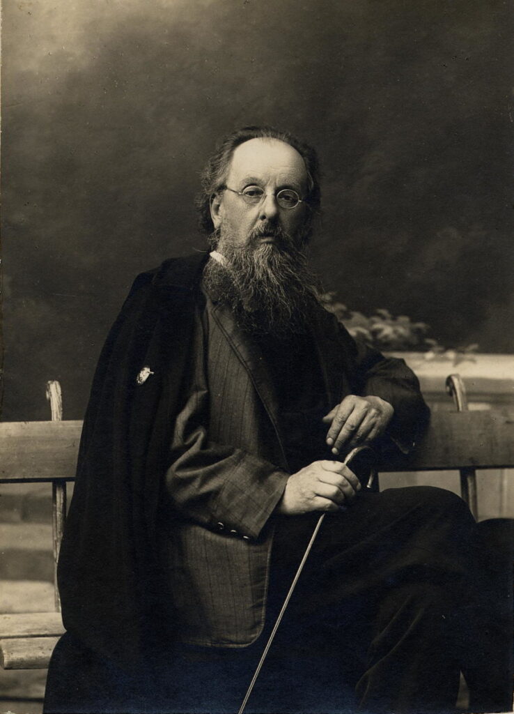 Konstantin Tsiolkovski, um homem idoso com uma longa barba branca e óculos redondos, está sentado em um banco. Ele segura uma bengala com as mãos, transmitindo uma postura serena e contemplativa. Considerado um dos pioneiros da astronáutica, suas ideias ajudaram a moldar o futuro das viagens espaciais.