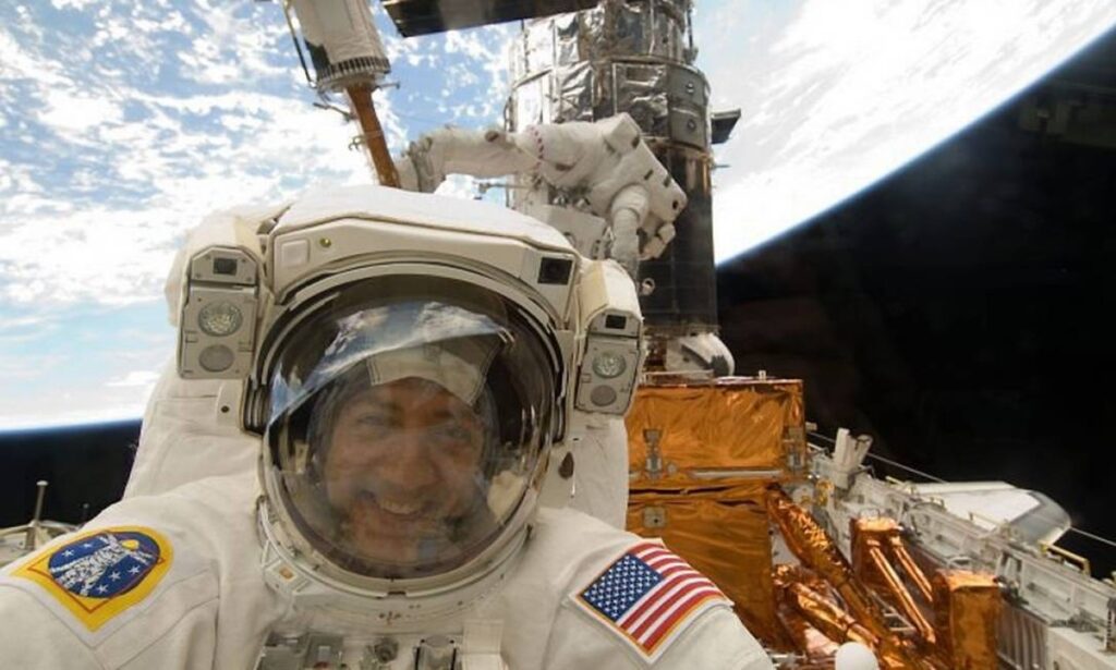 O astronauta Mike Massimino é fotografado do lado de fora da janela do ônibus espacial Atlantis durante uma missão de reparo no Telescópio Espacial Hubble, com a estrutura do telescópio parcialmente visível no fundo.