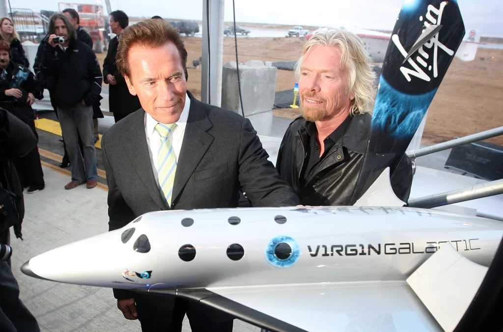 Richard Branson e Arnold Schwarzenegger em uma feira de tecnologia. Branson veste um blusão azul de estilo aeroespacial e está com as mãos atrás do corpo. Schwarzenegger está ao seu lado, ambos cercados por pessoas de terno. Próximos a eles, duas miniaturas de aviões da Virgin estão em exibição.
