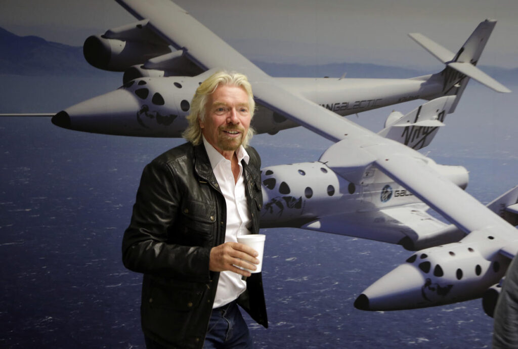 Richard Branson dando uma palestra, vestindo uma jaqueta preta. Ao fundo, há um banner na parede com a imagem e o nome da nave SS Unity, destacando o contexto aeroespacial do evento.