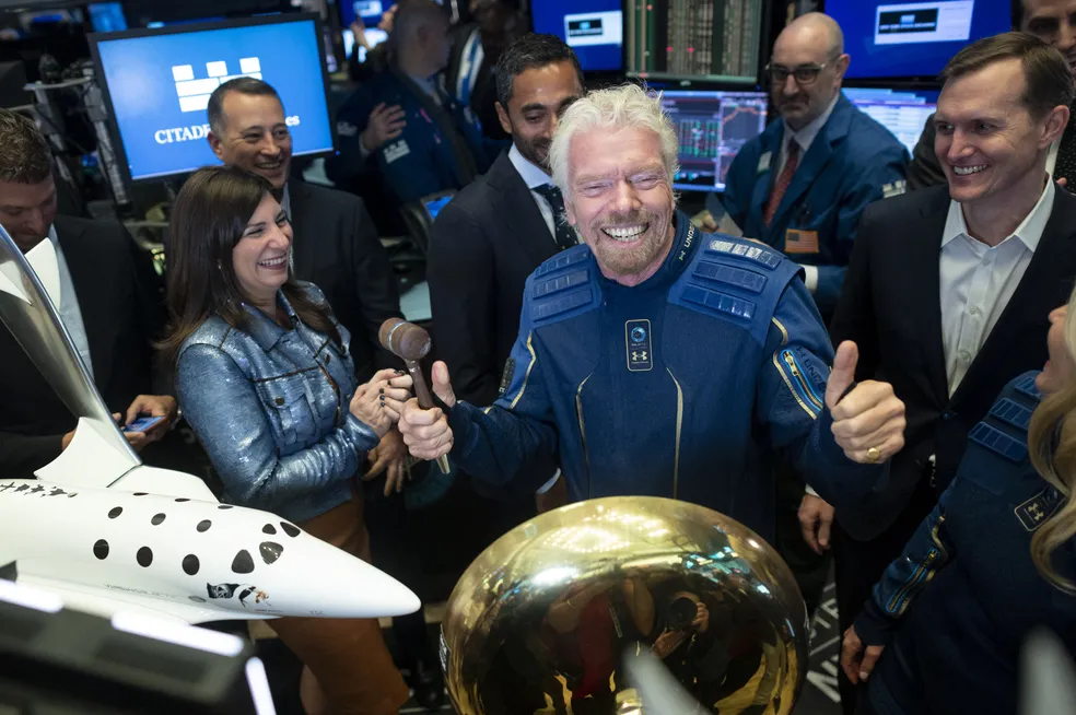 Richard Branson em uma feira de tecnologia, fazendo sinal de 'joia' com as duas mãos. Ele veste um blusão azul de estilo aeroespacial, rodeado por pessoas de terno. Ao lado dele, há uma miniatura de um avião da Virgin.