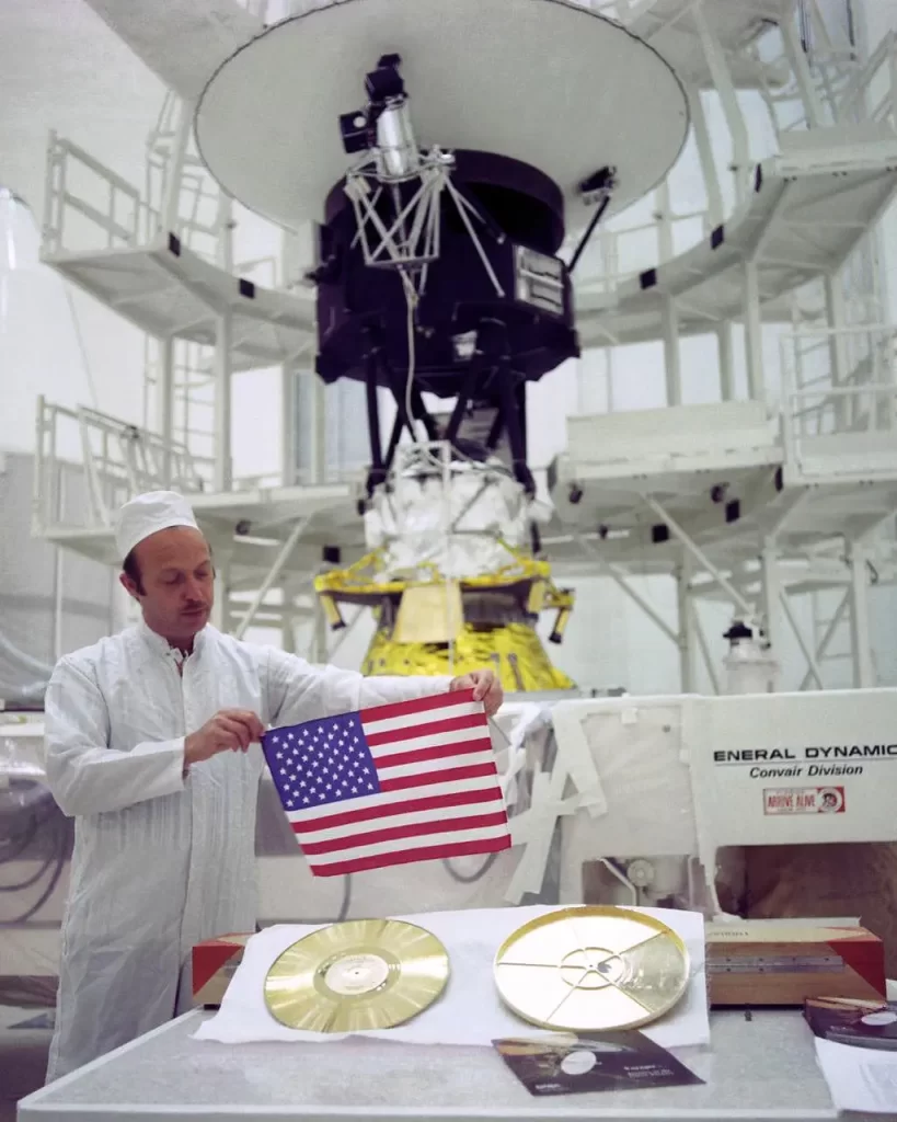 À medida que as duas espaçonaves Voyager da NASA viajam para o espaço profundo, elas carregam uma pequena bandeira americana e um Disco de Ouro repleto de imagens e sons — lembranças do nosso planeta natal. Esta foto mostra John Casani, gerente do projeto Voyager em 1977, segurando uma pequena bandeira de Dacron que foi dobrada e costurada nas mantas térmicas das espaçonaves Voyager antes de seu lançamento, há 36 anos. Abaixo dele estão o Disco de Ouro (à esquerda) e sua capa (à direita). Ao fundo, vê-se a Voyager 2 antes de seguir para a plataforma de lançamento. A foto foi tirada em Cabo Canaveral, Flórida, em 4 de agosto de 1977.Sondas Voyager da NASA e a contribuição para Astronomia.