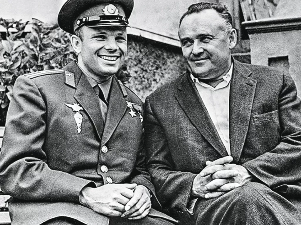 Yuri Gagarin, vestindo sua farda militar, e Sergei Korolev, usando um terno preto, estão lado a lado, ambos sorrindo. A cena captura um momento dos bastidores da Corrida Espacial, reunindo o primeiro cosmonauta da história e o brilhante engenheiro que esteve por trás do sucesso do programa espacial soviético.