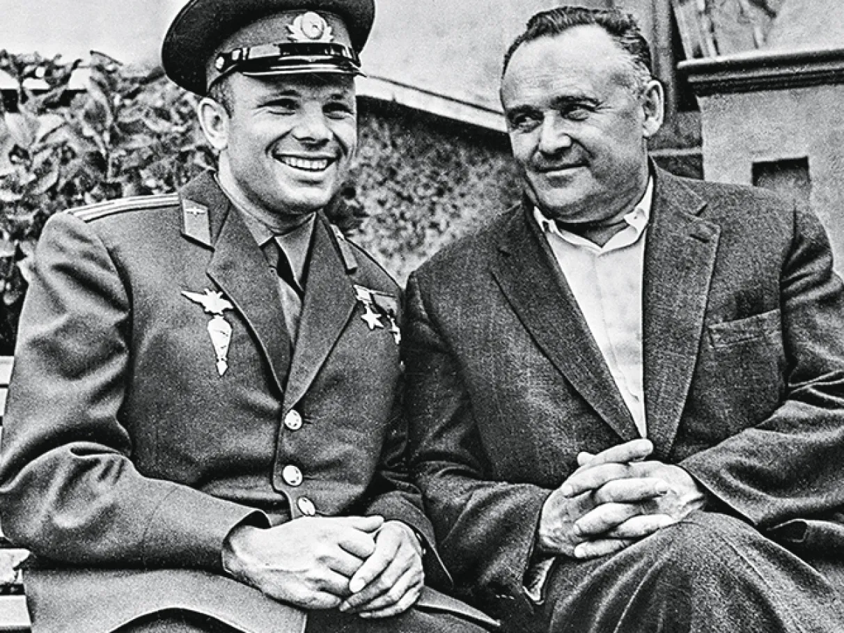 Yuri Gagarin, vestindo sua farda militar, e Sergei Korolev, usando um terno preto, estão lado a lado, ambos sorrindo. A cena captura um momento dos bastidores da Corrida Espacial, reunindo o primeiro cosmonauta da história e o brilhante engenheiro que esteve por trás do sucesso do programa espacial soviético.