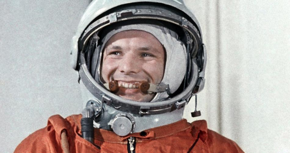 Yuri Gagarin, vestido com o traje espacial soviético laranja e um capacete branco com as letras "CCCP" na frente, exibe um sorriso radiante. Seu olhar transmite confiança e entusiasmo, refletindo o espírito pioneiro da primeira viagem tripulada ao espaço.