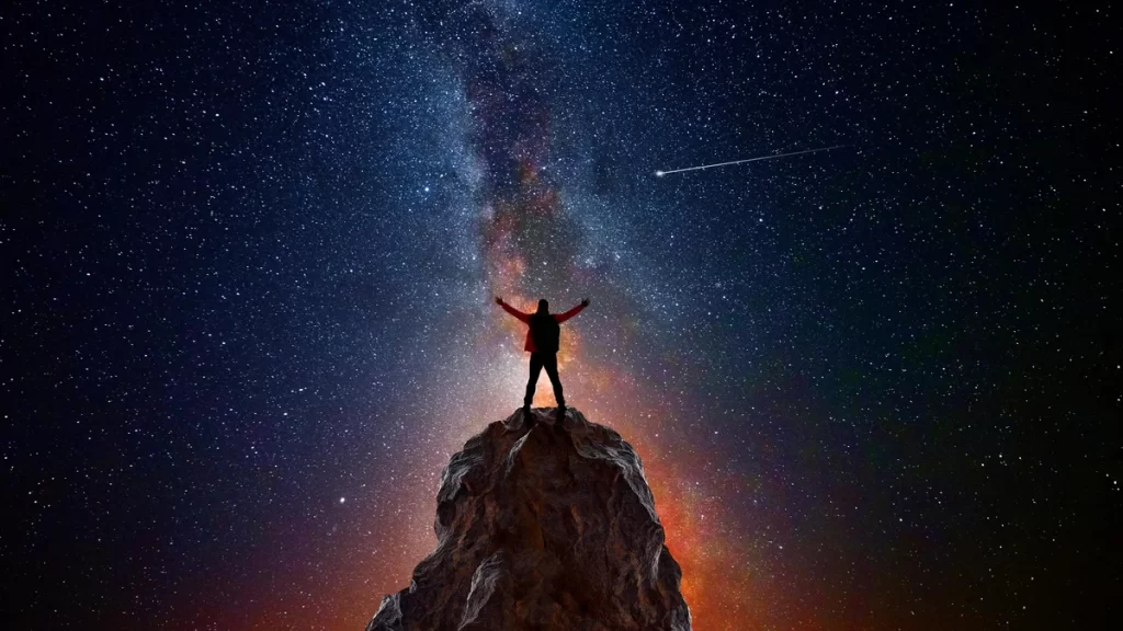Pessoa no topo de uma montanha com os braços levantados em direção ao céu noturno estrelado, observando um cometa e o braço da Via Láctea, representando como a astronomia define a conexão humana com o universo.