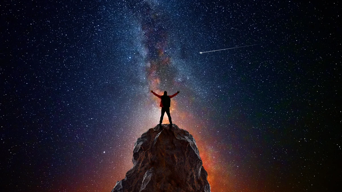Pessoa no topo de uma montanha com os braços levantados em direção ao céu noturno estrelado, observando um cometa e o braço da Via Láctea, representando como a astronomia define a conexão humana com o universo.