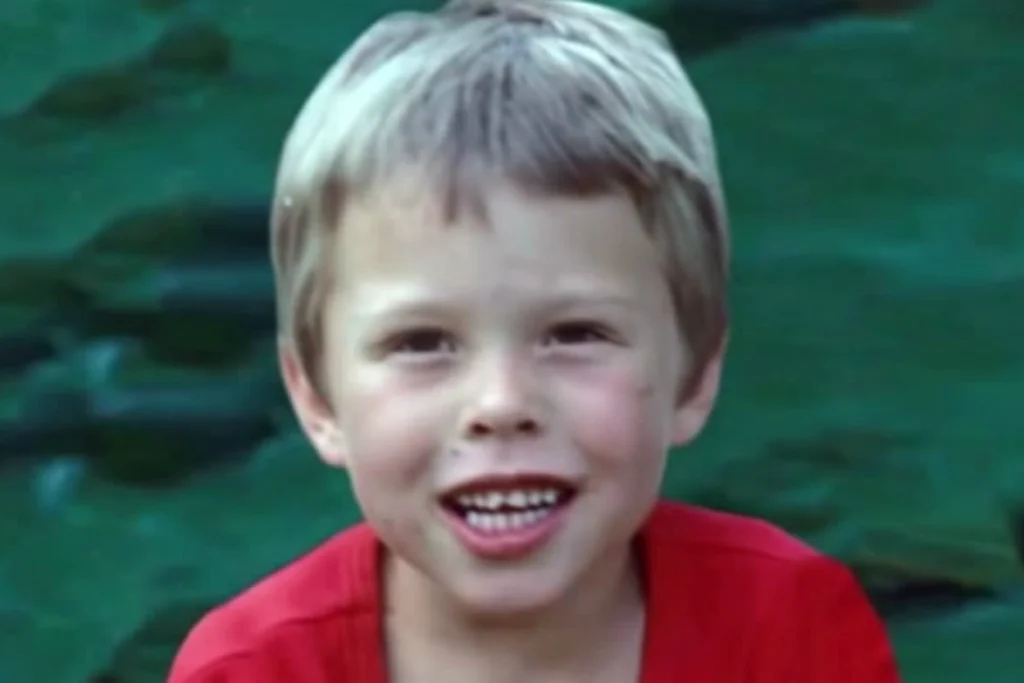 Elon Musk quando criança, vestindo uma camiseta vermelha, com sorriso tímido e dentes ainda em formação.