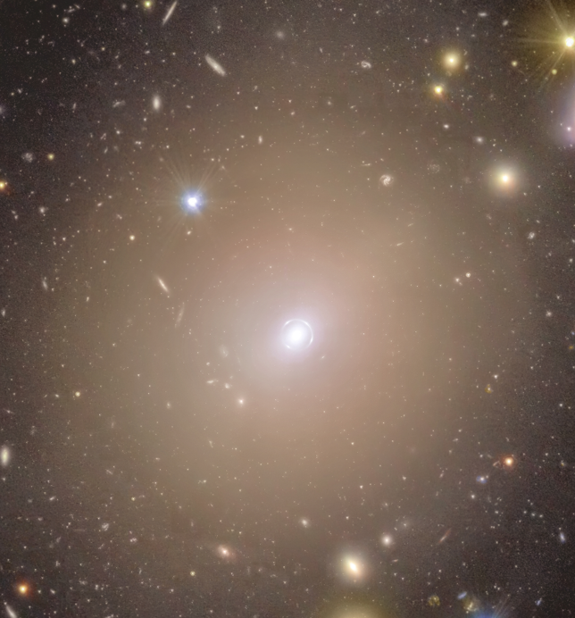 Foco no Anel Um mar de estrelas coloridas e galáxias parece nadar na imensidão escura do espaço ao redor de um halo nebuloso que ocupa o centro da cena. No meio da imagem, o aglomerado difuso de luz, em um tom quente de amarelo, se estende ao redor de um pequeno ponto brilhante, encaixado dentro de um círculo tênue de luz que parece traçado bem de perto ao seu redor. Conforme seguimos a borda do halo central para fora, seu brilho diminui e se mistura suavemente com o ambiente ao redor. Ali, discos alongados em tons que variam de um roxo quente a um dourado amarelado, além de pontos luminosos com pontas de difração bem definidas, se espalham de forma equilibrada por toda a imagem.