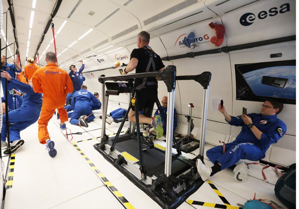 John McFall correndo na esteira durante um voo parabólico, usando camiseta e shorts pretos. Em um ambiente de microgravidade, ele está flutuando enquanto testa sua prótese de perna, com pilares ao fundo que fazem parte do treinamento. Seu foco está evidente enquanto mantém o ritmo na esteira, demonstrando a adaptação e o esforço físico necessários para o teste em condições de gravidade zero.