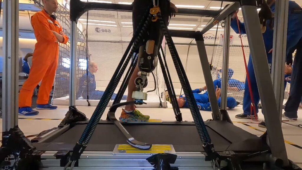 "Close na prótese de perna de John McFall enquanto ele corre na esteira durante um voo parabólico. A prótese é visível em detalhes, com foco na adaptação tecnológica para ambientes de microgravidade. Ao fundo, outros astronautas, vestidos com trajes espaciais azuis, flutuam enquanto observam o experimento, criando um contraste com a cena de teste da prótese em movimento.