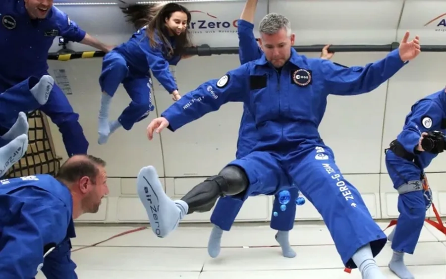 John McFall, o primeiro astronauta com deficiência física a testar prótese em microgravidade, veste um traje espacial azul enquanto realiza experimentos em um voo parabólico. Ele está flutuando ao lado de outros astronautas, todos em ambiente de microgravidade, durante uma sessão de treinamento. McFall é o único com uma prótese visível, demonstrando a adaptação tecnológica para deficientes físicos em ambientes espaciais.