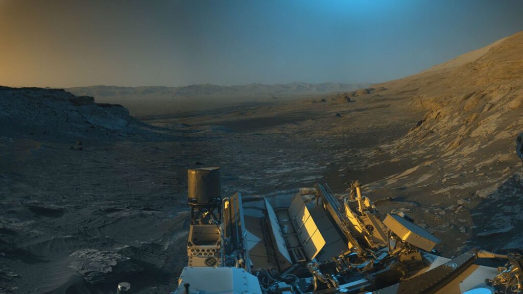 O rover Curiosity da NASA em Marte usou suas câmeras de navegação em preto e branco para capturar panoramas desta cena em dois momentos do dia. As cores azul, laranja e verde foram adicionadas a uma combinação dos dois panoramas para uma interpretação artística da cena.