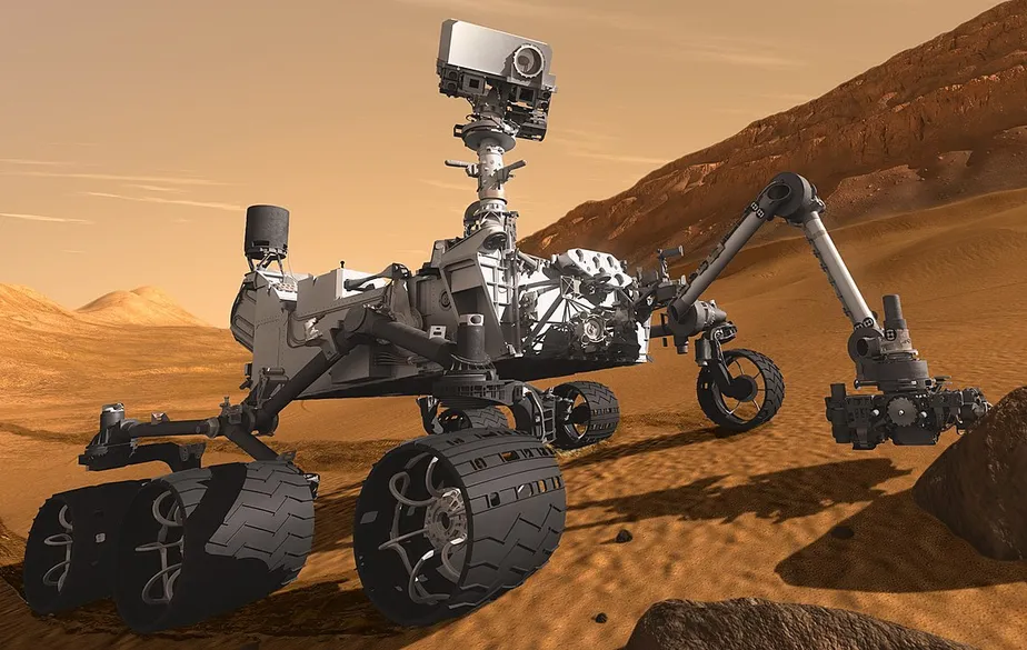 Representação artística do rover Curiosity da NASA em Marte, mostrando o veículo robótico de seis rodas sobre uma superfície rochosa avermelhada, com colinas e o céu marciano em tons alaranjados ao fundo. O rover aparece equipado com seu mastro de câmeras levantado, seus painéis e instrumentos cobertos por poeira marciana, enquanto realiza exploração científica da paisagem ao redor.