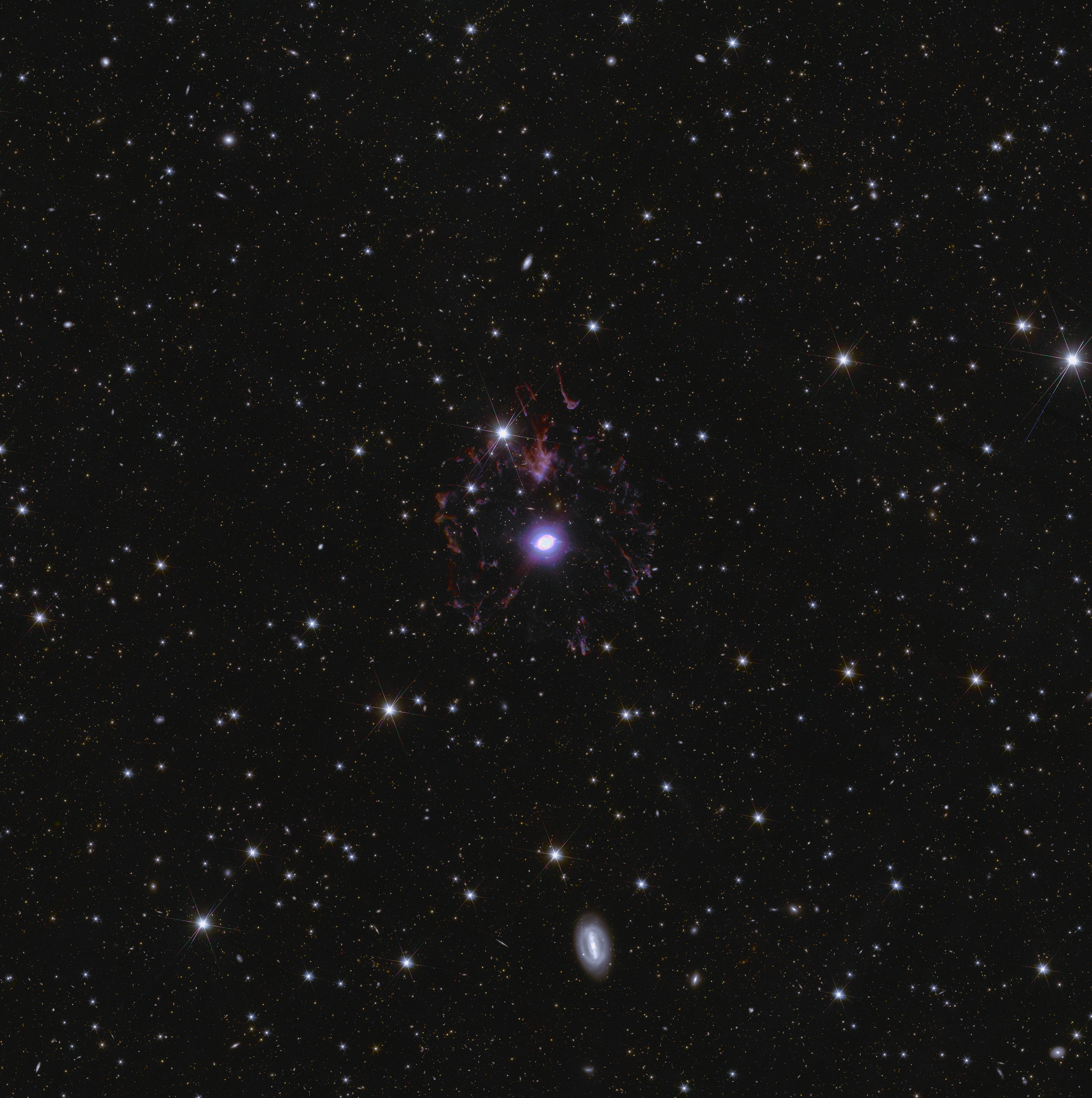 A Nebulosa do Olho de Gato ocupa o centro do palco em um mar cintilante de estrelas e galáxias. Um ponto está localizado no centro da nebulosa — é a visão da estrela moribunda. Ao seu redor, há uma série de camadas e laços complexos e coloridos de gás e poeira que foram ejetados pela estrela ao longo do tempo. Mais afastados, há filamentos e manchas de gás e poeira em várias formas e tamanhos, que dão a impressão de fragmentos de um balão estourado congelados ao redor do ponto de explosão. Milhões de galáxias estão ao fundo. Numerosas estrelas brilhantes com distintos picos de difração também são claramente visíveis.