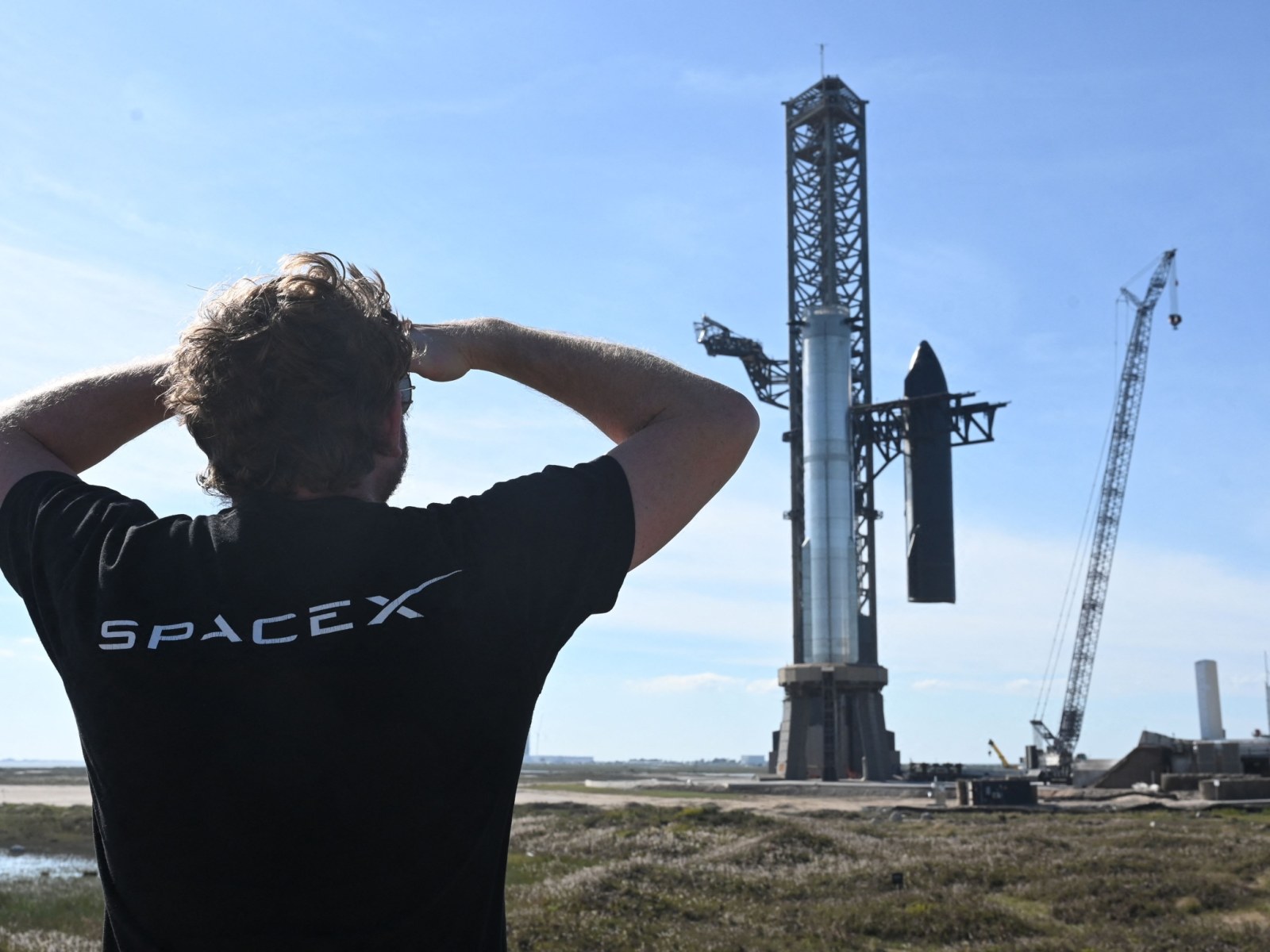 Captura do Super Shuper heavy Elon Musk de costas, vestindo uma camiseta preta com o logo da SpaceX, observa o Super Heavy e a Starship sendo segurados pelos chopsticks do Mechazilla.