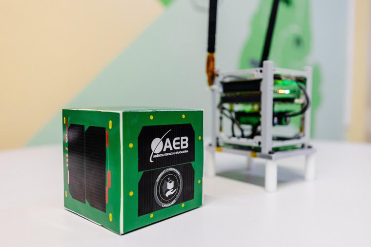 Imagem do nanossatélite brasileiro Aldebaran‑1, em formato CubeSat 1U, com acabamento verde e logotipo da Agência Espacial Brasileira (AEB) visível. Ao fundo, bancada de testes com estrutura eletrônica e antena acoplada, representando o processo de validação tecnológica antes do lançamento orbital.