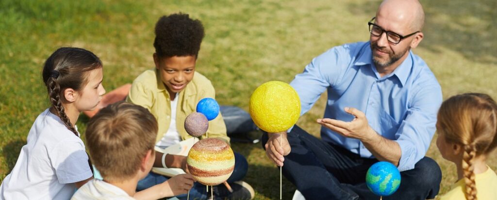  Professor ensinando astronomia nas escolas ao ar livre para um grupo de crianças, usando maquetes dos planetas do sistema solar para explicar conceitos do espaço.