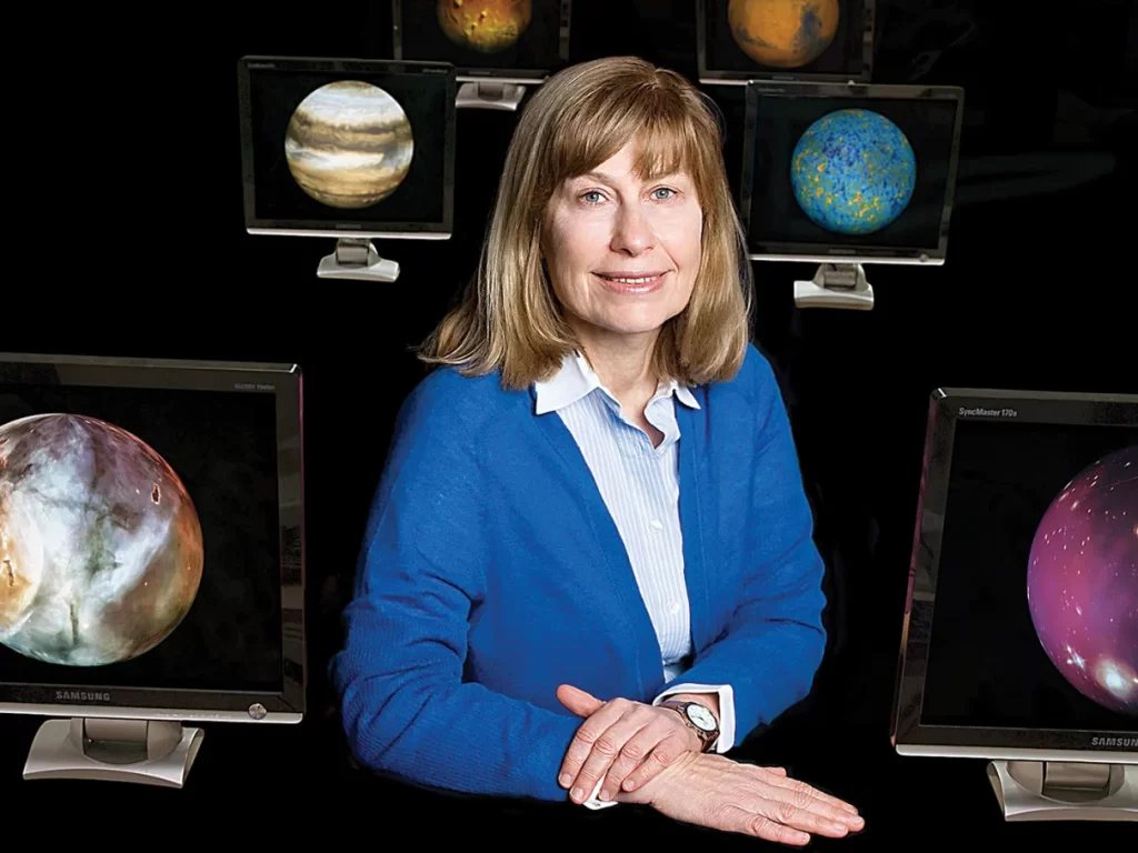 Beatriz Barbuy sorrindo em seu ambiente de trabalho, cercada por vários monitores que mostram imagens de planetas, enquanto analisa dados astronômicos.