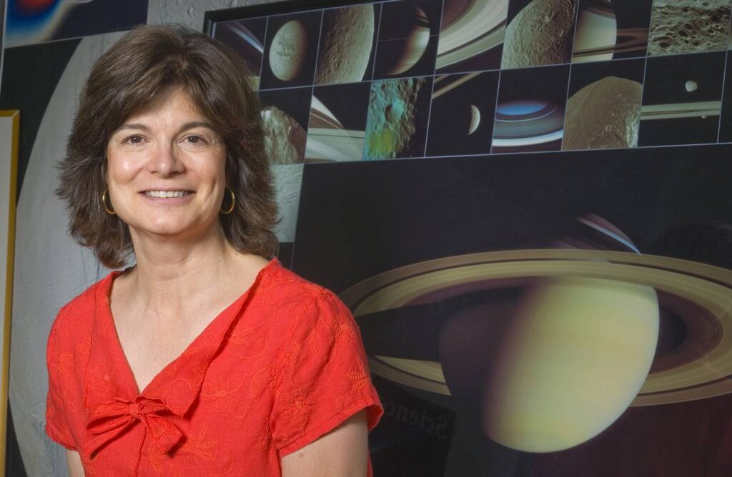 Imagem de Carolyn Porco, astrônoma planetária, usando camiseta vermelha e sorrindo, com imagens de Saturno e seus anéis ao fundo.