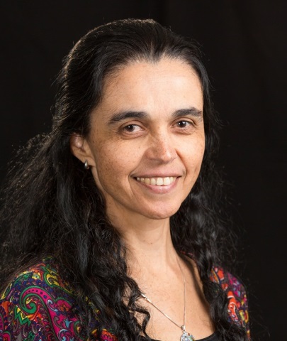 Claudia Lucia Mendes de Oliveira sorrindo, com cabelo preto solto, usando brincos e um vestido colorido, em um ambiente relacionado à astronomia.
