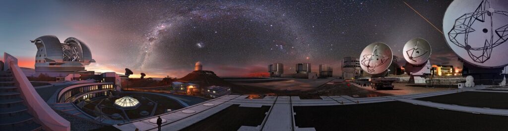 Panorama noturno do Observatório Europeu do Sul (ESO), no deserto do Atacama, mostrando telescópios e radiotelescópios sob o céu estrelado e a Via Láctea ao fundo. Além disso, as instalações do Very Large Telescope e do ALMA aparecem iluminadas, compondo uma vista espetacular do centro de observação astronômica.