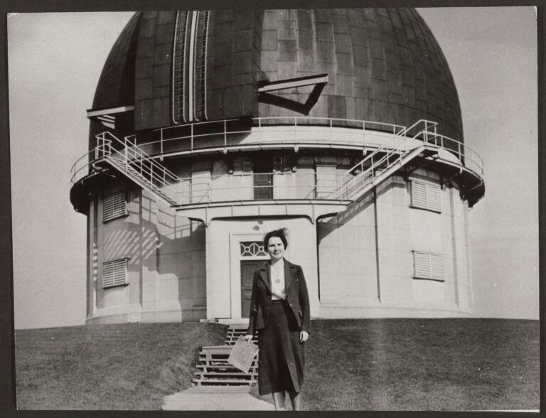 Foto em preto e branco de Helen Sawyer Hogg, importante astrônoma e divulgadora científica, usando roupa de época em frente à cúpula de um observatório astronômico.