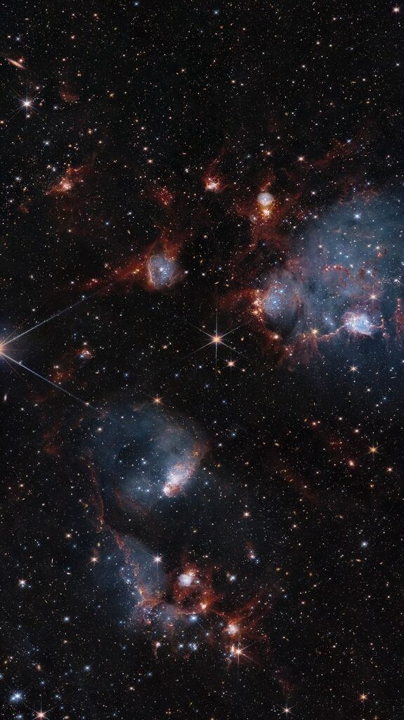 Nuvens azuladas de gás estão entrelaçadas com faixas e filamentos de poeira brilhando em vermelho.
Essas nuvens contêm cavidades repletas de estrelas, e muitas outras estrelas são visíveis ao fundo e espalhadas por entre as nuvens.
As concentrações de nuvens se destacam na parte superior direita e no centro inferior da imagem.
Pequenas galáxias de fundo também estão salpicadas por toda a cena.