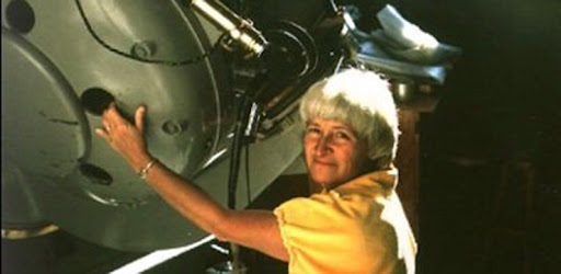 Imagem de Carolyn Shoemaker, astrônoma e caçadora de cometas, com cabelos brancos, usando camiseta amarela, sorrindo levemente na frente de um telescópio, com fotografias de cometas e imagens espaciais ao fundo.