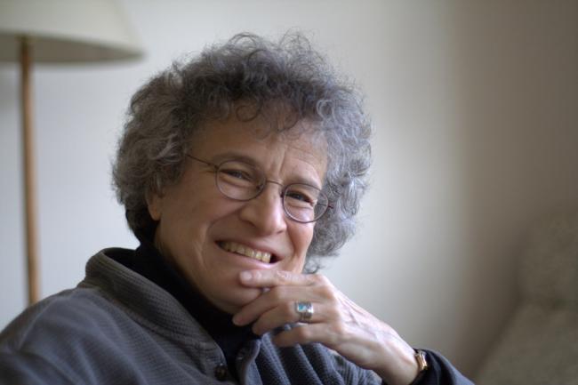 Imagem de Margaret Geller, astrônoma renomada, com cabelo grisalho, óculos e sorriso acolhedor, em ambiente científico.