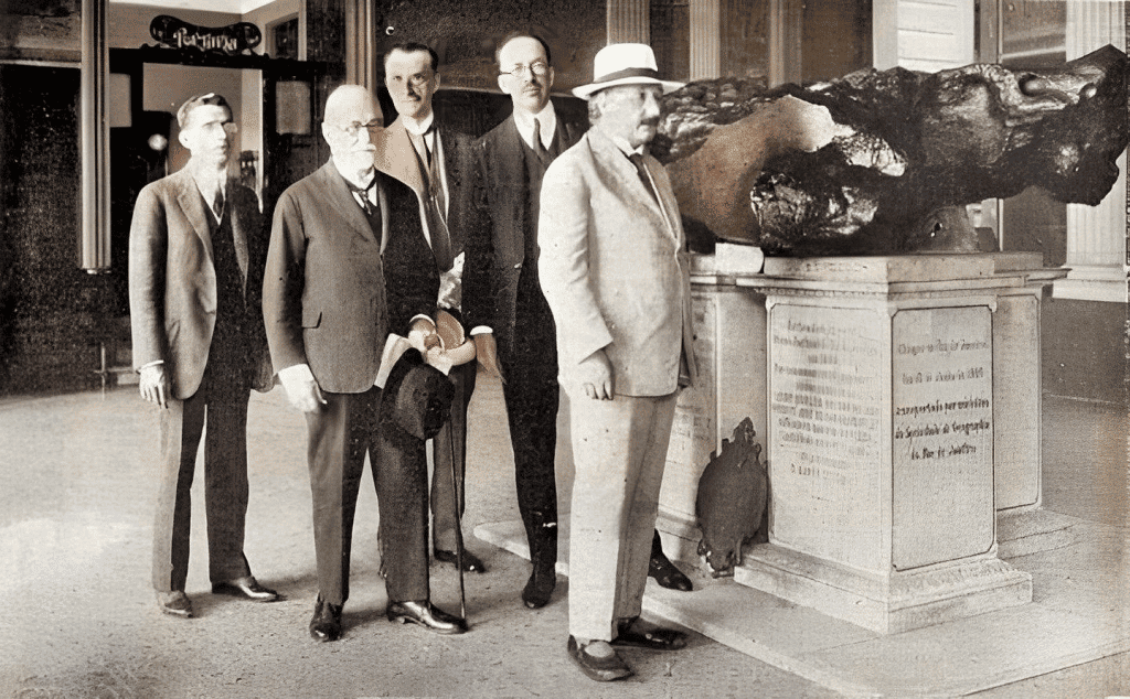 Fotografia histórica de 1925 mostrando um grupo de homens, incluindo Albert Einstein, em visita ao meteorito Bendegó no Museu Nacional do Rio de Janeiro. O meteorito está exposto sobre pedestais com inscrições descritivas.
