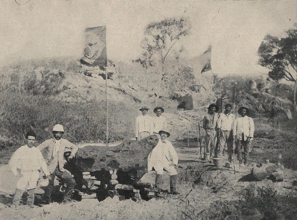 Meteorito de Bendegó em fotografia de H. Antunes, tirada em 1887 mostrando o meteorito ainda na margem do riacho Bendegó,com o vice-almirante José Carlos de Carvalho e dos engenheiros Humberto Saraiva Antunes e Vicente José de Carvalho. Ao fundo, tremula a Bandeira do Império do Brasil.