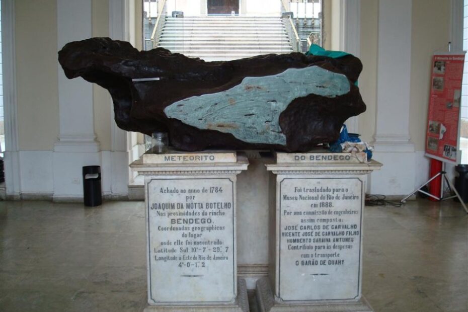 Fotografia do meteorito Bendegó exposto no Museu Nacional do Rio de Janeiro. A peça, composta por ferro e níquel, está apoiada sobre dois pedestais de mármore com inscrições históricas sobre sua descoberta em 1784 na Bahia e transporte para o museu em 1888.