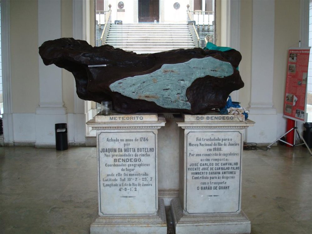 Fotografia do meteorito Bendegó exposto no Museu Nacional do Rio de Janeiro. A peça, composta por ferro e níquel, está apoiada sobre dois pedestais de mármore com inscrições históricas sobre sua descoberta em 1784 na Bahia e transporte para o museu em 1888.