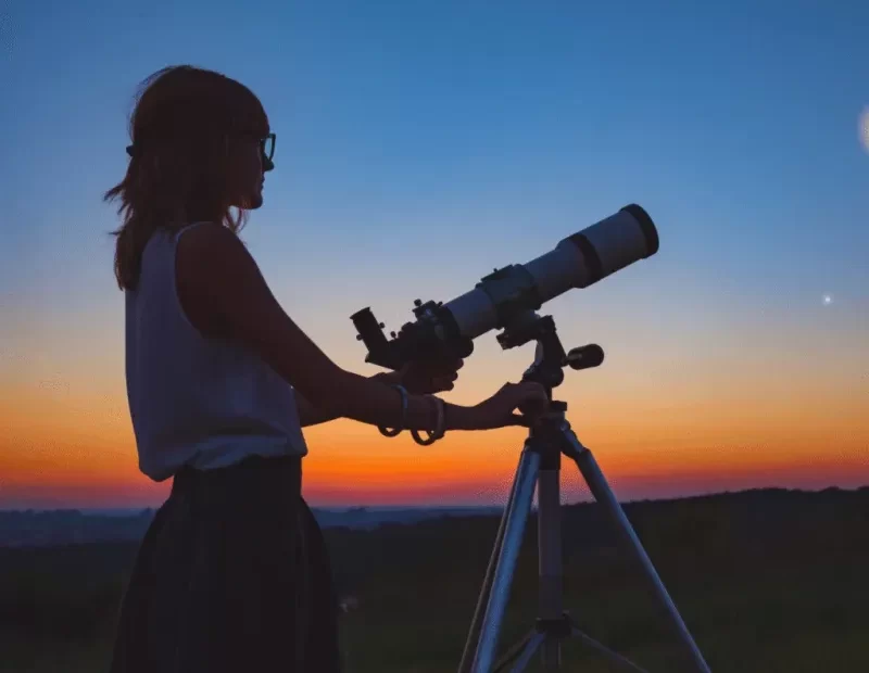 Mulher astrônoma com o seu telescópio ao fundo um céu com cores azul e alaranjado como se fosse o entardecer