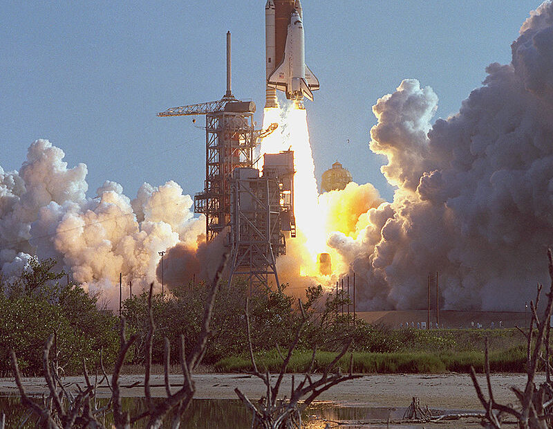 Ônibus espacial Discovery durante seu primeiro lançamento em 30 de agosto de 1984, deixando uma trilha de fumaça ao decolar da plataforma, com o céu claro ao fundo e estruturas da base de lançamento visíveis.