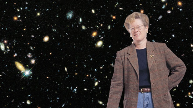 Imagem de Sandra Faber, astrônoma especialista em galáxias, usando calça jeans, terno marrom e sorriso simpático, com imagens de galáxias ao fundo.