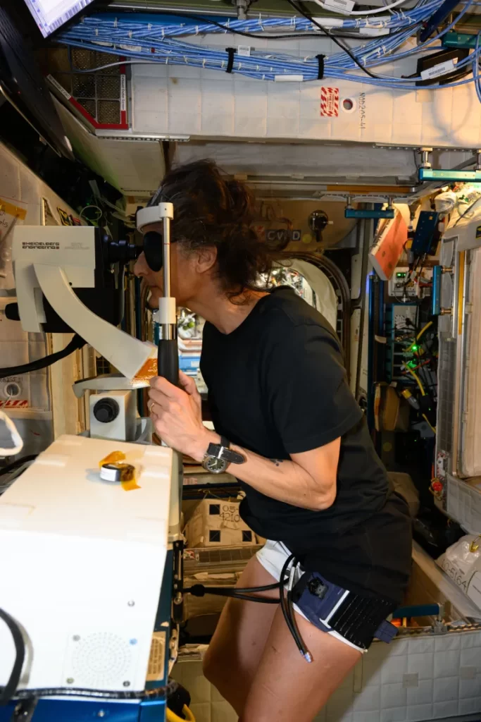 "Williams, vestindo uma camiseta preta, shorts brancos e meias pretas, está de frente para um equipamento semelhante às máquinas usadas por oftalmologistas na Terra. Ela usa uma braçadeira azul e preta na coxa esquerda, de onde saem dois cabos. Com a mão direita, segura uma barra do equipamento. Visão no espaço