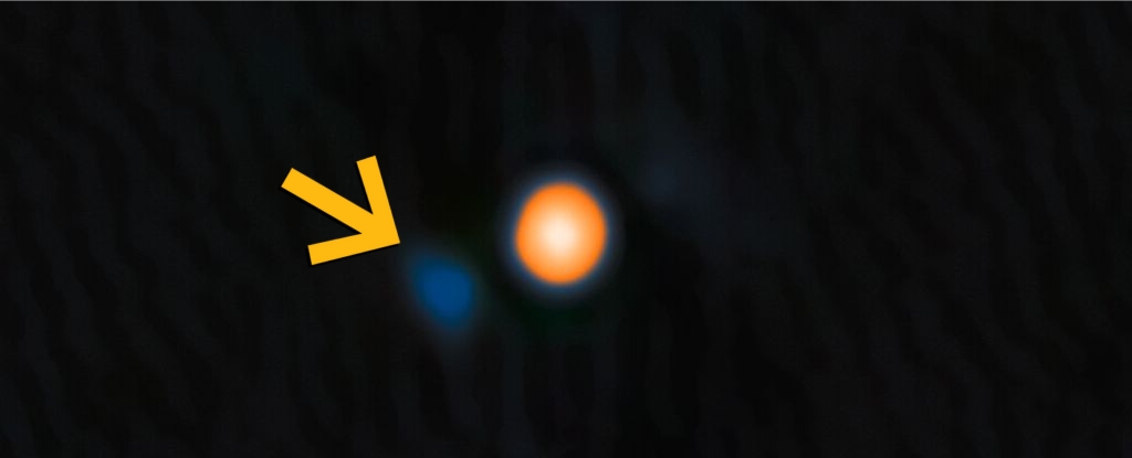 Imagem da estrela Betelgeuse em tons amarelo-avermelhados. Uma seta amarela destaca sua estrela companheira pr&oacute;xima, vis&iacute;vel como um ponto azul t&ecirc;nue. Dados: NASA, JPL e NOIRLab; visualiza&ccedil;&atilde;o: NOIRLab.