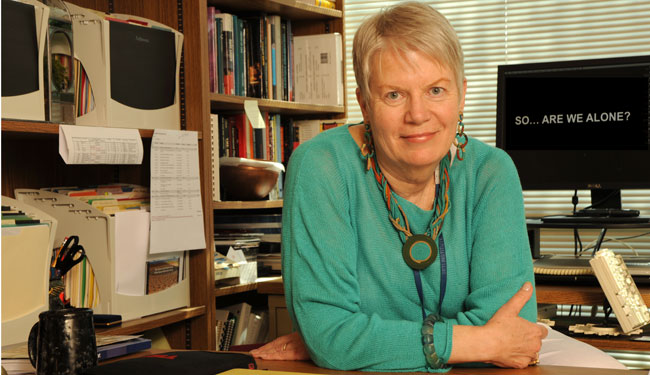 Imagem de Jill Tarter, astrônoma e pesquisadora do SETI, com cabelo curto e camisa verde, em uma sala com livros e estantes ao fundo.
