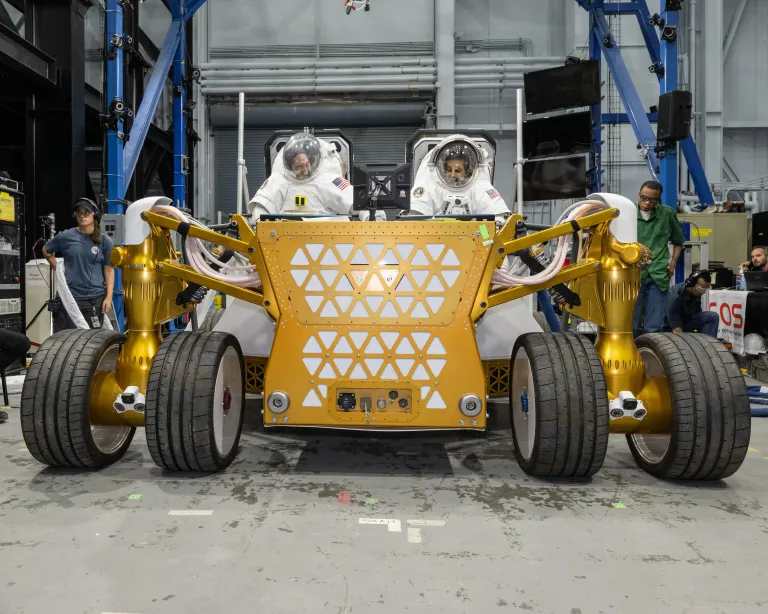 Engenheiros da NASA vestindo trajes espaciais sentados sobre um protótipo de rover lunar durante testes no Johnson Space Center, em Houston.