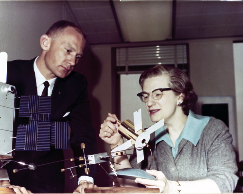 Alt text: Nancy Grace Roman, astrônoma pioneira da NASA, observa modelo de satélite ao lado de colega durante reunião técnica nos anos 1960. Conhecida como "Mãe do Hubble", Roman foi fundamental para o desenvolvimento do Telescópio Espacial Hubble.