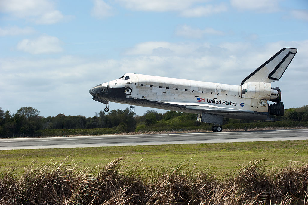 Ônibus espacial Discovery pousando na Terra durante sua última missão, STS-133, em 9 de março de 2011, com fumaça visível nas rodas tocando a pista após o contato, marcando o fim de sua trajetória histórica.