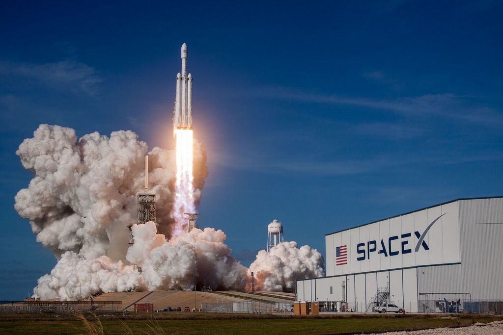 Foguete Falcon Heavy da SpaceX decolando da base de lançamento sob céu azul claro, com fumaça branca na base