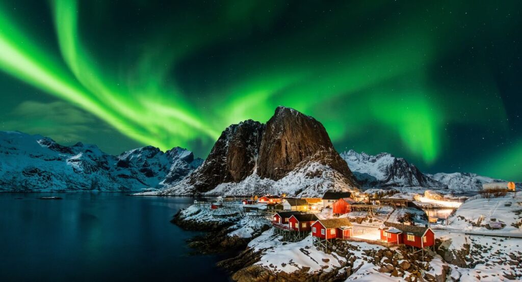 Aurora boreal iluminando o céu noturno sobre uma casa típica da Noruega, com luzes verdes e roxas dançando acima da paisagem nevada.