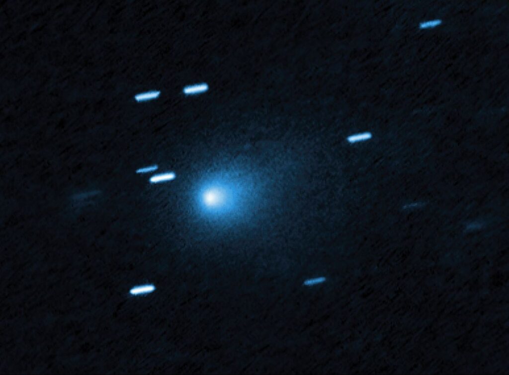 No centro da imagem está um cometa que se apresenta como um casulo azulado em formato de gota, formado por poeira que se desprende do núcleo sólido e gelado do cometa, visto contra um fundo preto. O cometa parece estar se movendo em direção ao canto inferior esquerdo da imagem. Cerca de uma dúzia de rastros diagonais curtos e azul-claro estão espalhados pela imagem — são estrelas de fundo que pareceram se mover durante a exposição, já que o telescópio estava acompanhando o movimento do cometa.