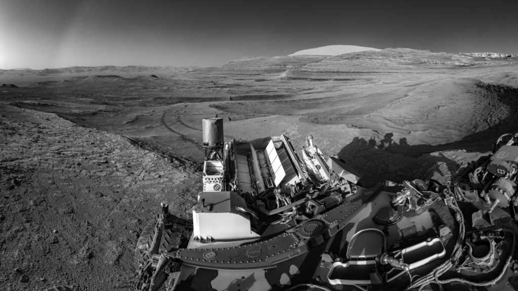 Nesta imagem registrada em 26 de julho de 2025, é possível ver as trilhas deixadas pelo rover Curiosity da NASA enquanto ele transmitia dados simultaneamente para um orbitador em Marte. Essa combinação de tarefas demonstra uma estratégia eficiente de uso da energia gerada por sua fonte nuclear — visível ao fundo, com fileiras de aletas brancas.